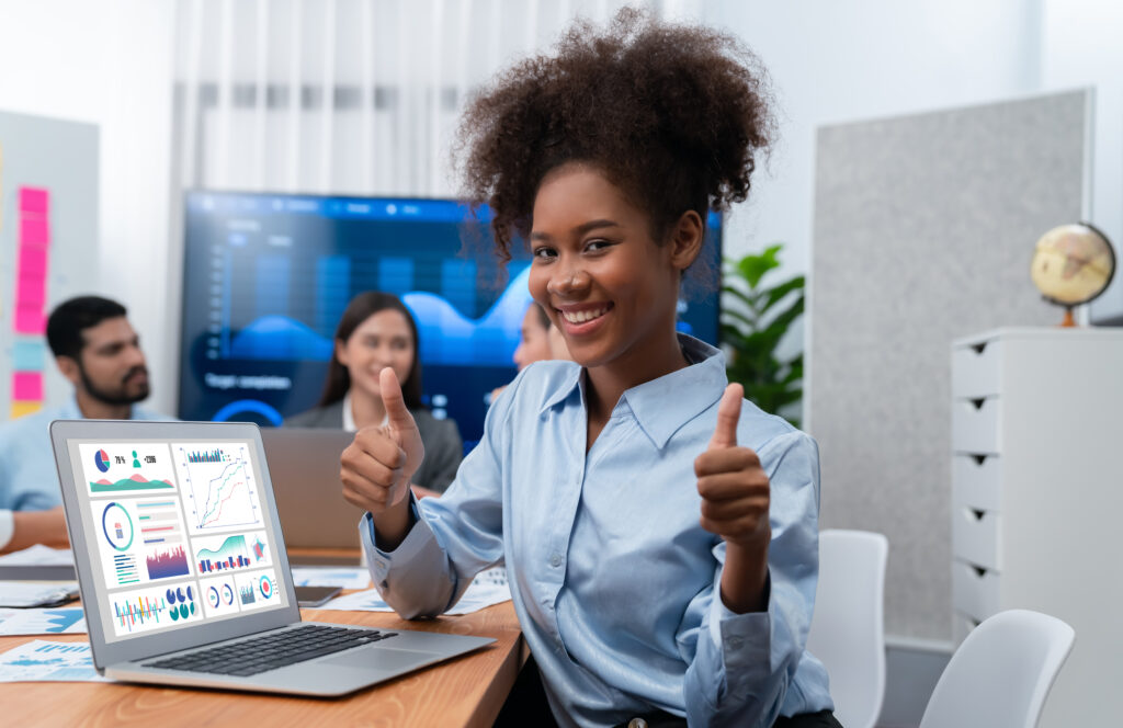 Portrait of happy young african businesswoman with group of office worker on meeting with screen display business dashboard in background. Confident office lady at team meeting. Concord