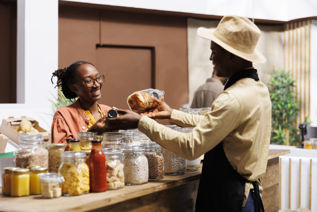 African American woman shops at a local supermarket, discussing organic, bulk produce with friendly shopkeeper. They focus on plastic-free, sustainable options, and promoting a zero-waste lifestyle.